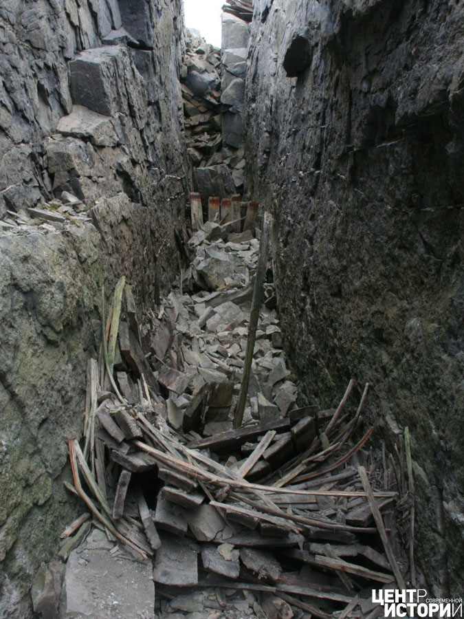 В узкой расщелине, среди глыб и щебня, виден выступающий кусочек доски